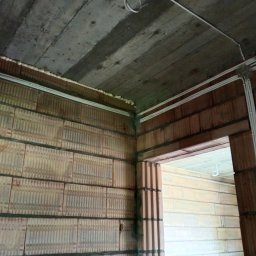 Jakub Ogar - Surowy mur z pustaków ceramicznych z widocznymi poziomymi kanałami instalacyjnymi, zabezpieczonymi pianką montażową przy suficie, oraz białe korytka kablowe prowadzone wzdłuż ścian i nadproża...