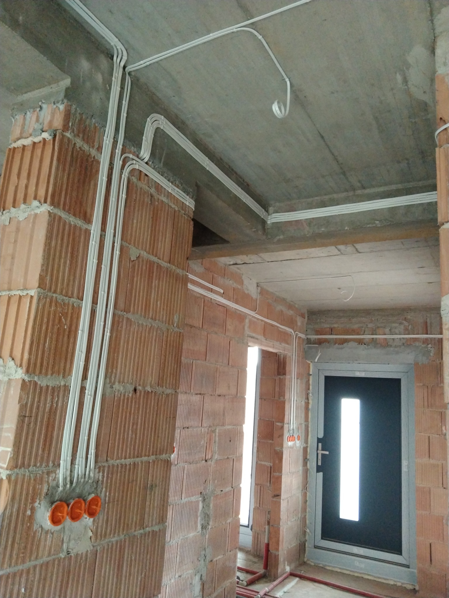 Instalacja elektryczna natynkowa w surowym wnętrzu: białe kable prowadzone po ceglanej ścianie i betonowym suficie, widoczne pomarańczowe puszki instalacyjne i czarne drzwi wejściowe z długim...