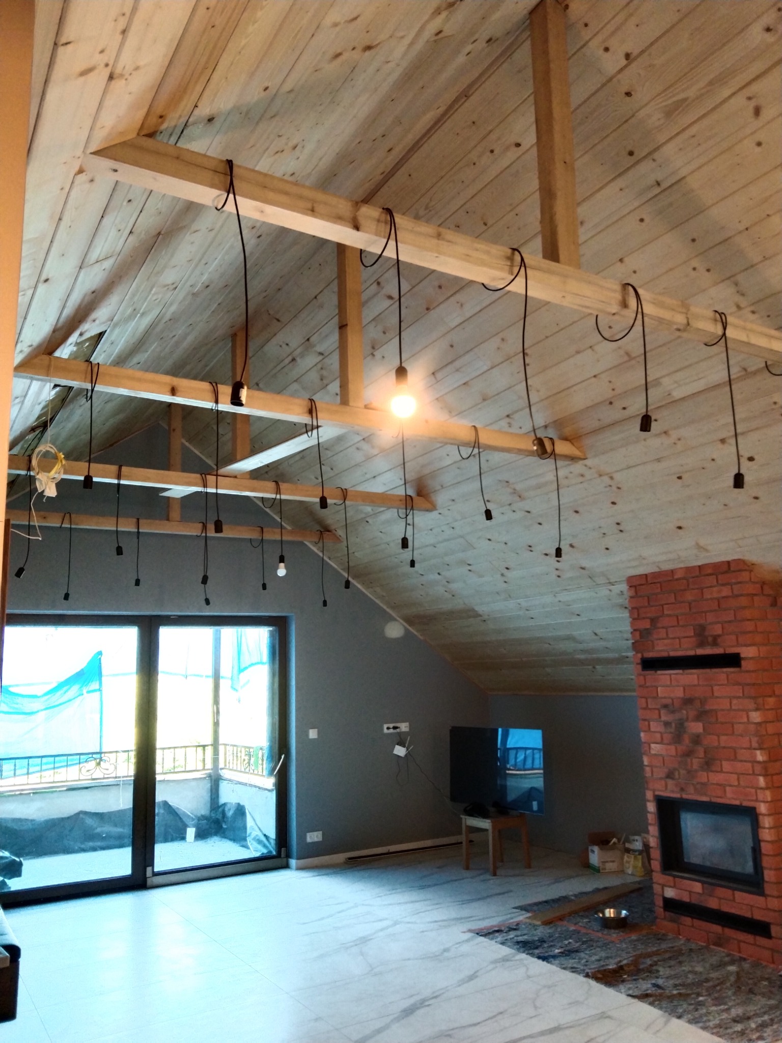 Wnętrze poddasza z drewnianym stropem i widocznymi belkami konstrukcyjnymi, na których zawieszone są przewody z oprawkami żarówek, jedna żarówka świeci, przygotowanie do montażu oświetlenia, w tle...