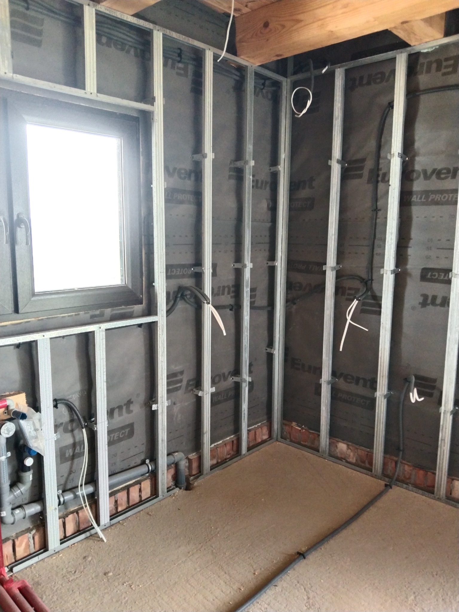 Wnętrze w trakcie remontu: metalowa konstrukcja szkieletowa ścian, okno z czarną ramą, przewody elektryczne wystające ze ściany, częściowo widoczne rury hydrauliczne przy podłodze.