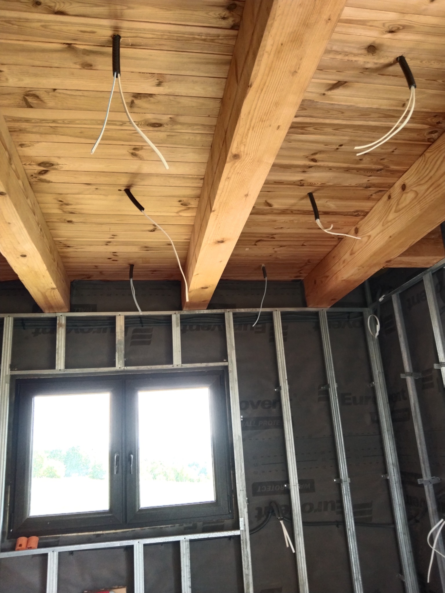 Surowy stan wnętrza: drewniany sufit z widocznymi belkami, z którego zwisają przewody w peszlach; ściany w trakcie budowy z widoczną konstrukcją metalową i oknem.