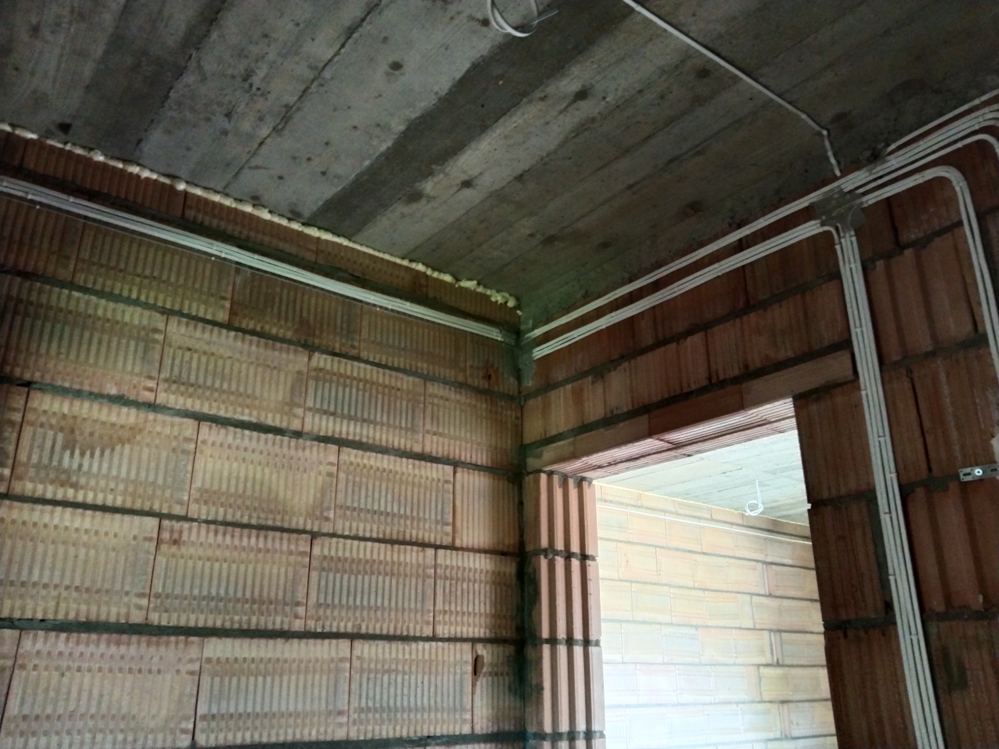 Surowy mur z pustaków ceramicznych z widocznymi poziomymi kanałami instalacyjnymi, zabezpieczonymi pianką montażową przy suficie, oraz białe korytka kablowe prowadzone wzdłuż ścian i nadproża...