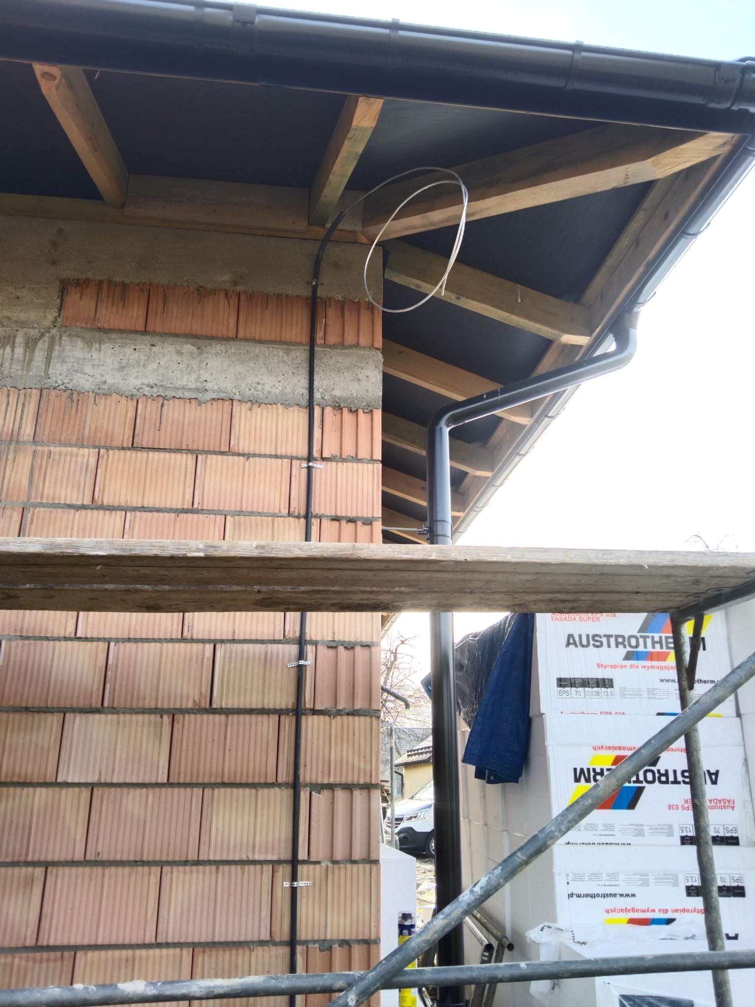 Czarny kabel elektryczny przymocowany do ściany z cegły klinkierowej na zewnątrz budynku, widoczny fragment dachu z rynną i rusztowanie z materiałami budowlanymi w tle.