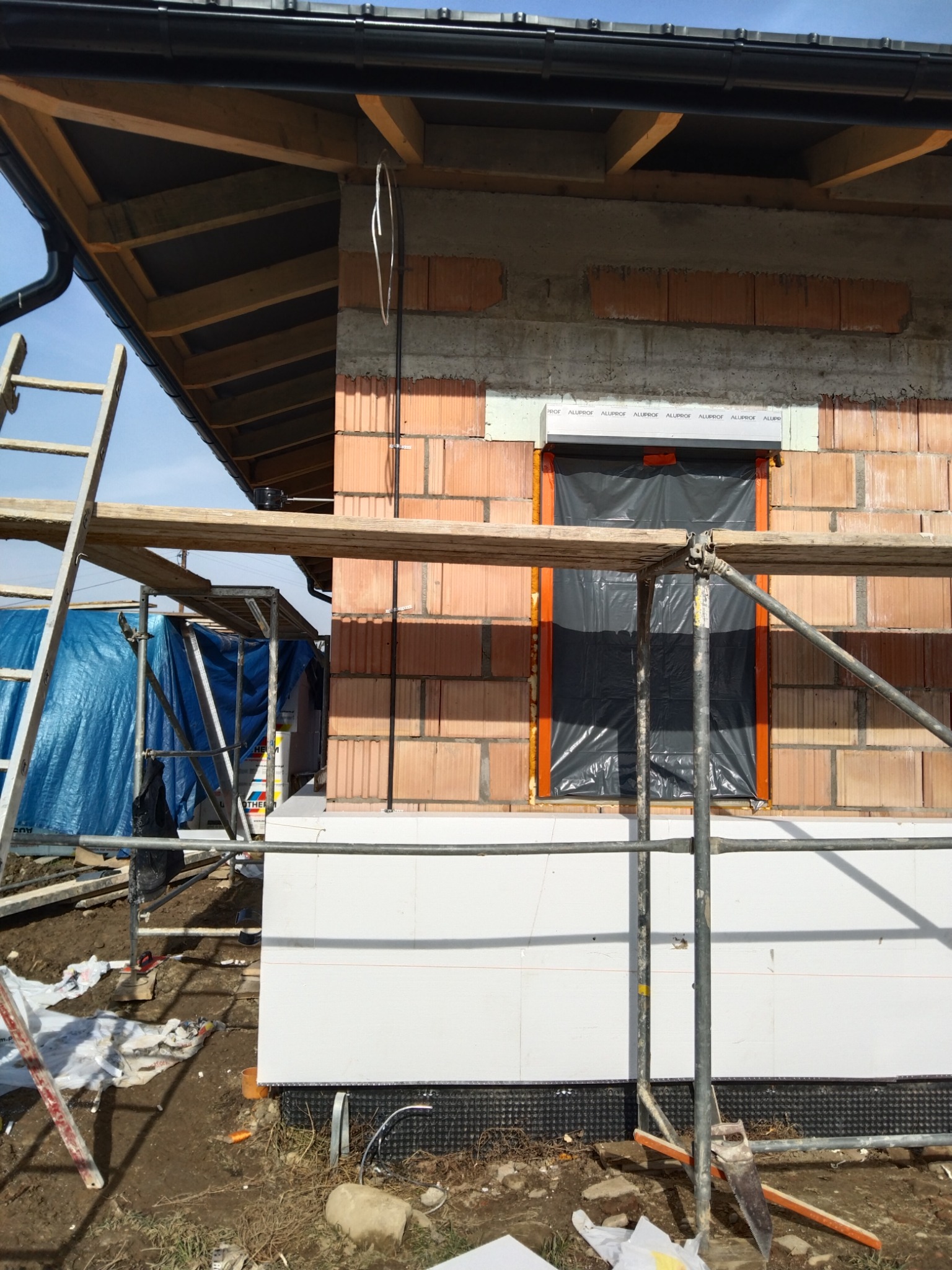 Budynek w trakcie budowy z widocznymi przewodami elektrycznymi na ścianie z cegły, rusztowaniem i oknem zabezpieczonym folią.