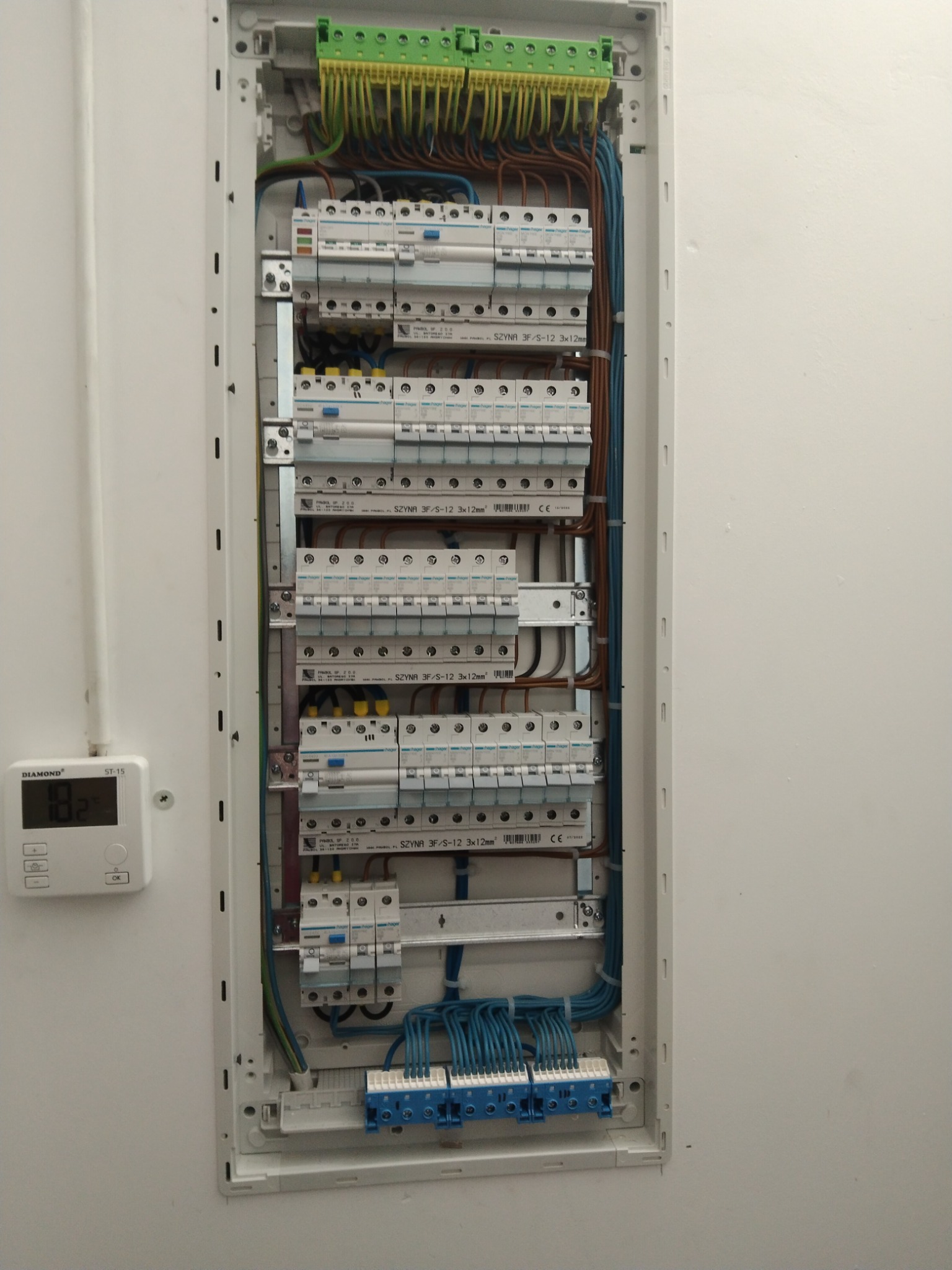 Otwarta skrzynka elektryczna z widocznymi bezpiecznikami, listwami zaciskowymi i przewodami w różnych kolorach, umieszczona na białej ścianie obok termostatu pokazującego temperaturę 18.2 stopnia...