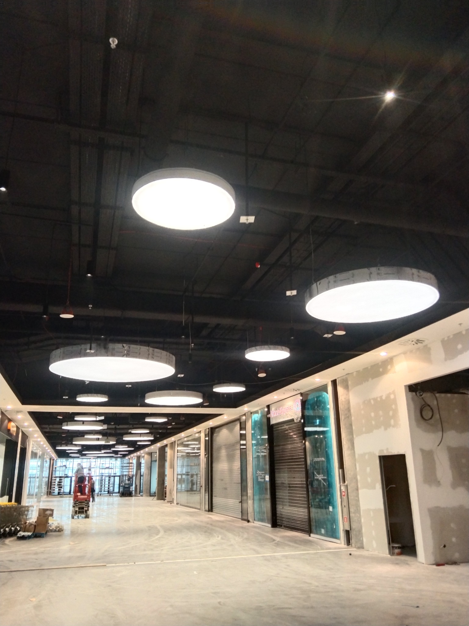Montaż okrągłych lamp sufitowych w nowym centrum handlowym, widoczne instalacje elektryczne i sufit podwieszany.