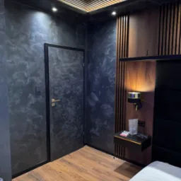 Pokój z ciemnymi ścianami o marmurkowym wzorze, drewnianą podłogą, drewnianą zabudową ściany z lampką nocną i podwieszanym sufitem z drewnianymi listwami.