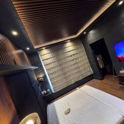 Gbel-bud - Nowoczesna sypialnia z drewnianym sufitem lamelowym, czarnymi ścianami i dużym oknem z roletą rzymską. Na podłodze panele, a w głębi widoczne wejście do garderoby.