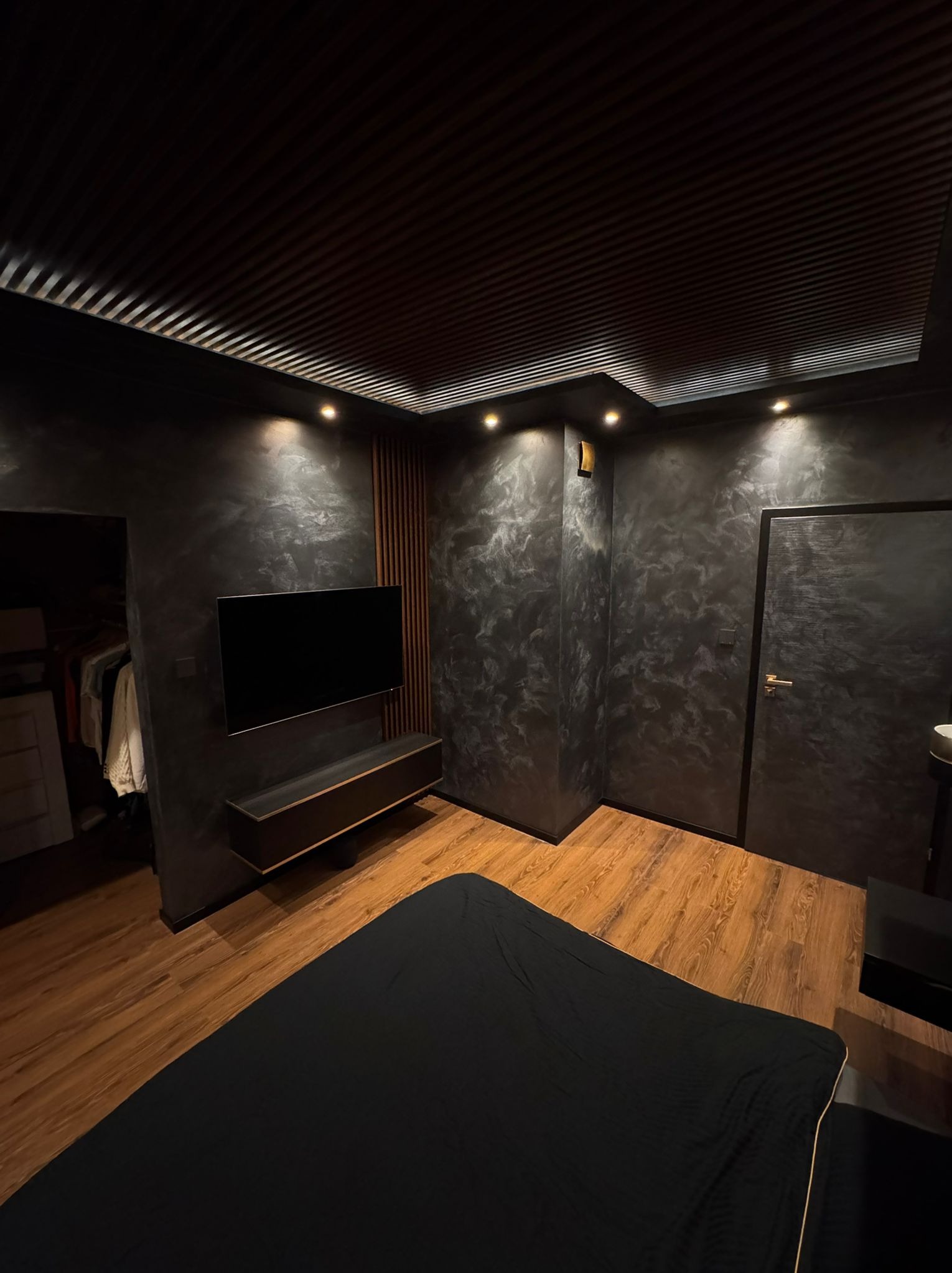 Sypialnia z czarnymi ścianami wykończonymi efektem dekoracyjnym, drewnianą podłogą, podwieszanym sufitem z lamelami i wpuszczanymi lampami, telewizorem na ścianie i minimalistyczną szafką RTV