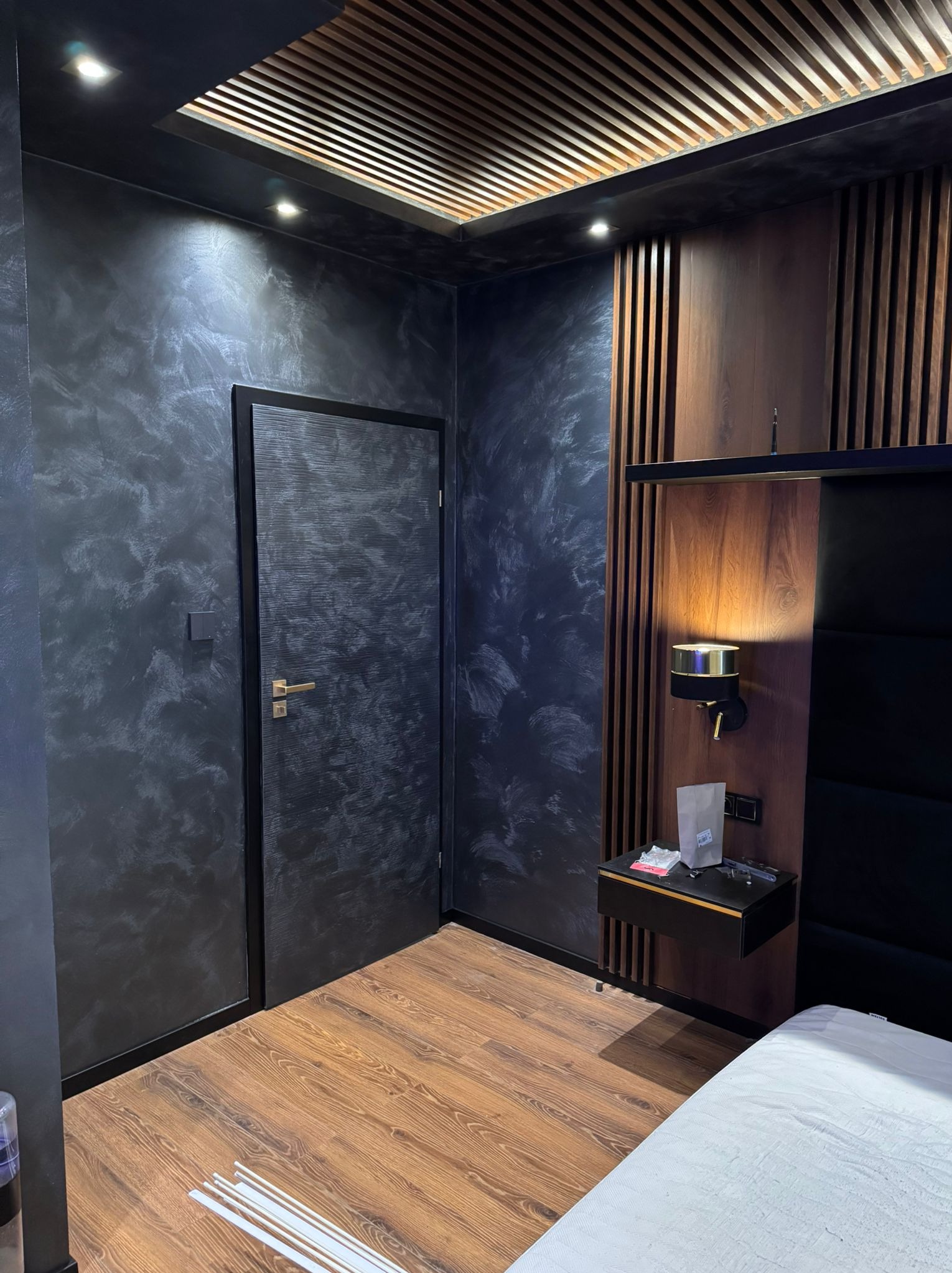 Pokój z ciemnymi ścianami o marmurkowym wzorze, drewnianą podłogą, drewnianą zabudową ściany z lampką nocną i podwieszanym sufitem z drewnianymi listwami.
