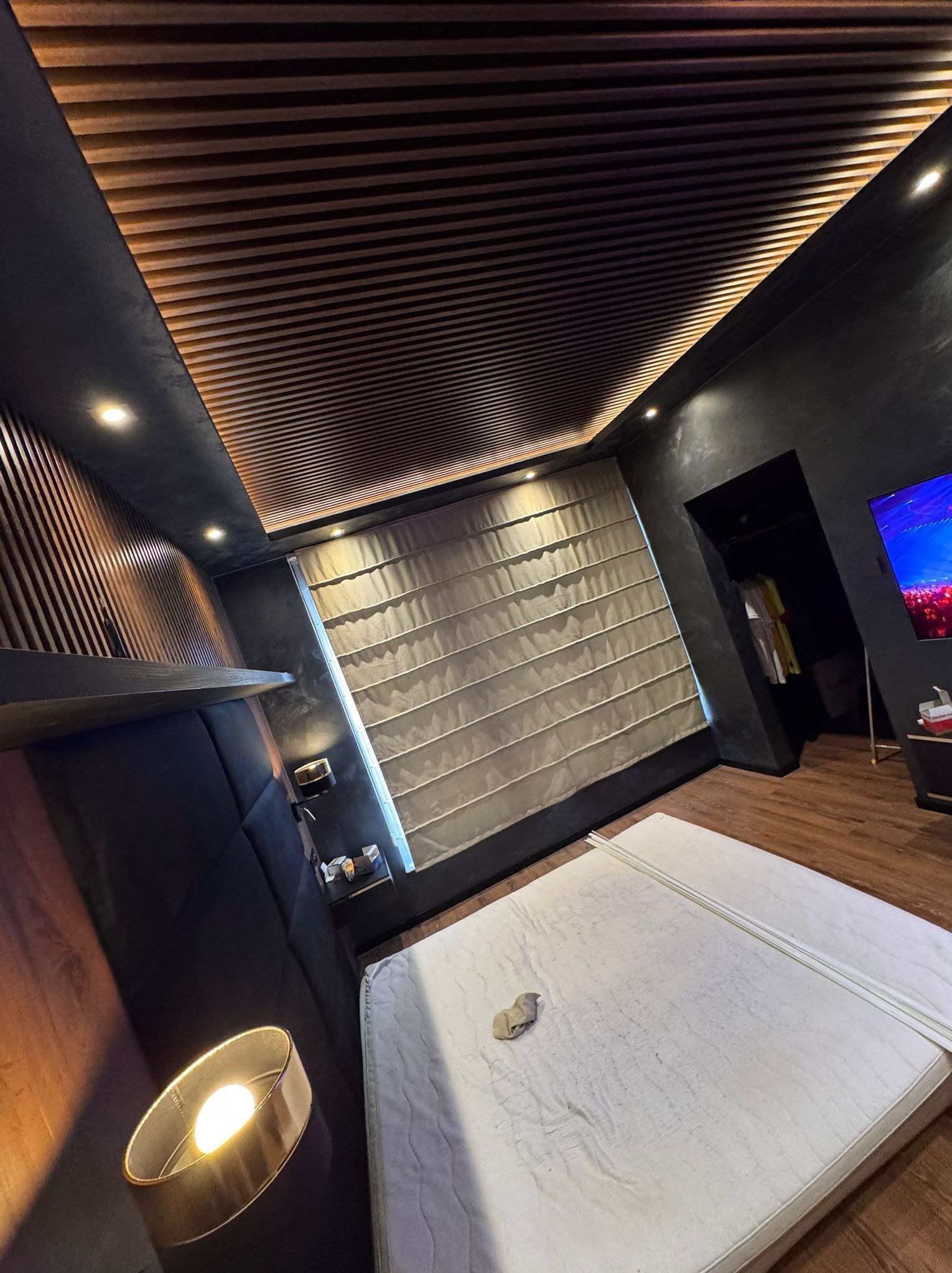 Nowoczesna sypialnia z drewnianym sufitem lamelowym, czarnymi ścianami i dużym oknem z roletą rzymską. Na podłodze panele, a w głębi widoczne wejście do garderoby.