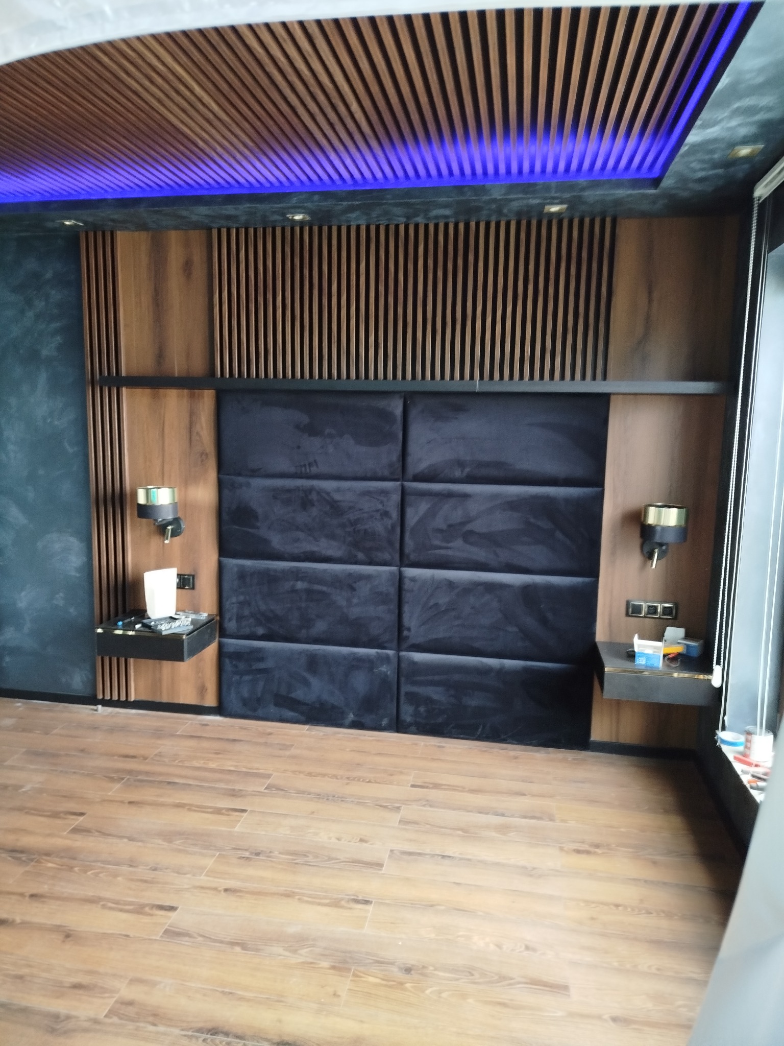 Nowoczesna sypialnia z drewnianymi lamelami na suficie i ścianie, podświetleniem LED, tapicerowanym panelem zagłówka z czarnego weluru i drewnianą podłogą imitującą deski.