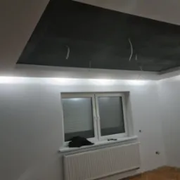 Pokój po remoncie: białe ściany, podświetlany sufit podwieszany z ciemną wnęką, widoczne wystające kable, okno z żaluzjami i grzejnik.