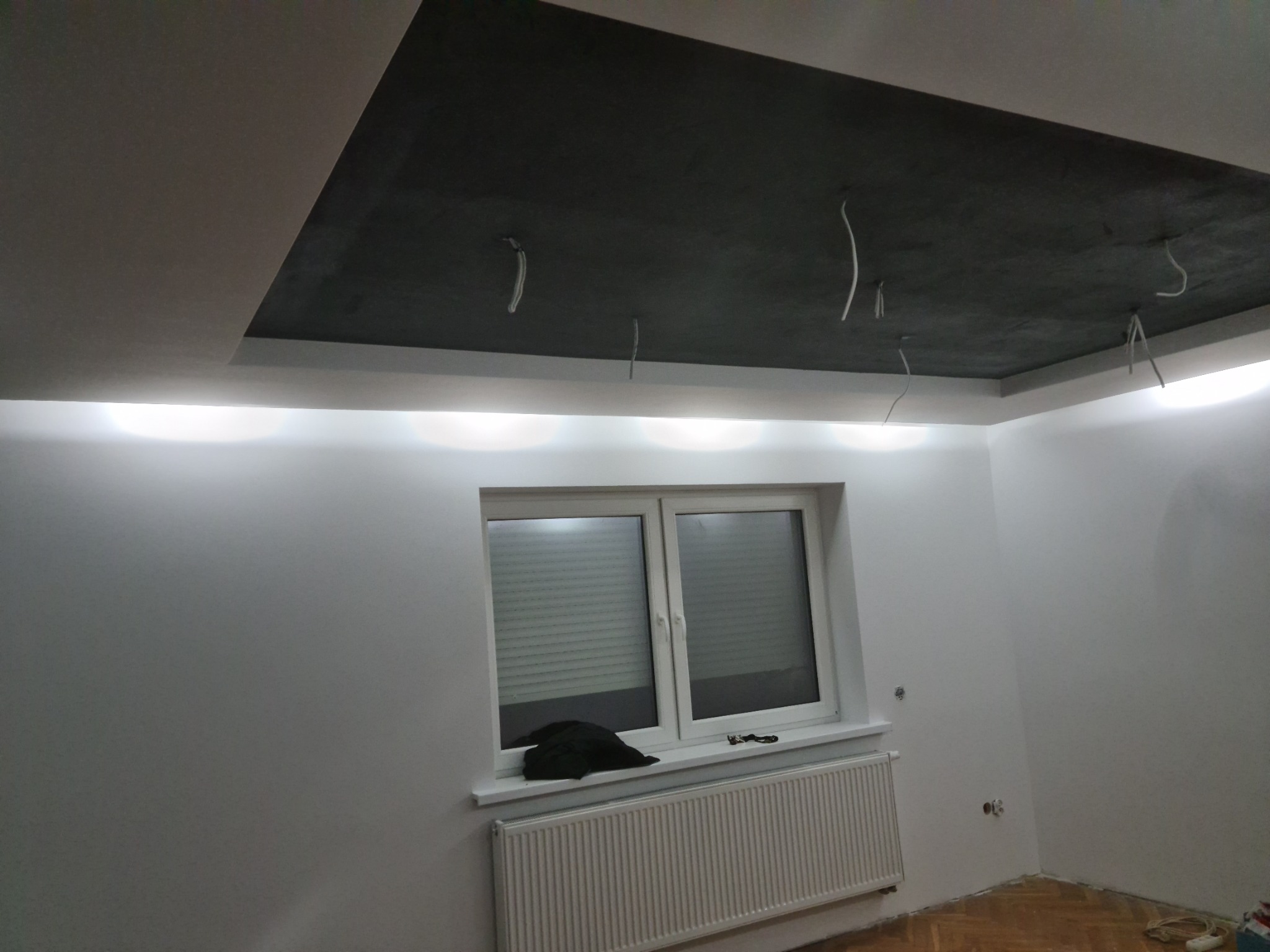 Pokój po remoncie: białe ściany, podświetlany sufit podwieszany z ciemną wnęką, widoczne wystające kable, okno z żaluzjami i grzejnik.