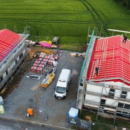 INSTEP PLUS SPÓŁKA Z OGRANICZONĄ ODPOWIEDZIALNOŚCIĄ - Budowa bliźniaków z paneli CLT z czerwonym pokryciem dachowym i białymi ścianami. Widok z góry na plac budowy z materiałami i samochodem dostawczym.