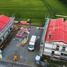 Budowa bliźniaków z paneli CLT z czerwonym pokryciem dachowym i białymi ścianami. Widok z góry na plac budowy z materiałami i samochodem dostawczym.