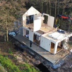 Montaż domu szkieletowego z drewnianych paneli, widok z góry na plac budowy z dźwigiem i pracownikami w lesie.