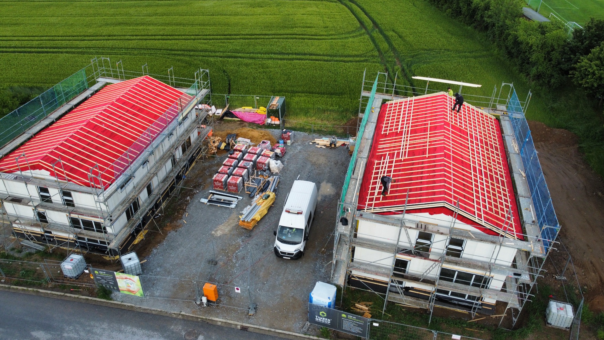 Budowa bliźniaków z paneli CLT z czerwonym pokryciem dachowym i białymi ścianami. Widok z góry na plac budowy z materiałami i samochodem dostawczym.