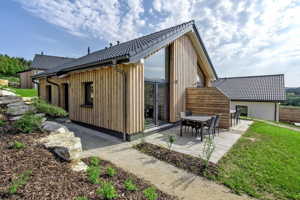 Nowoczesny dom z elewacją z jasnego drewna, ciemną dachówką i dużymi oknami, z tarasem i ogrodem.