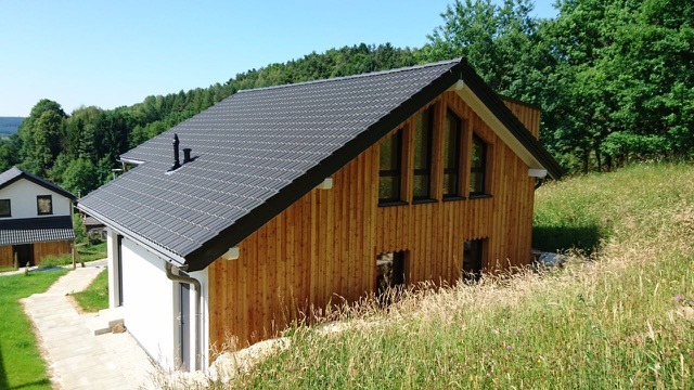 Nowoczesny dom z drewnianą elewacją i grafitowym dachem, usytuowany na zboczu porośniętym trawą, z widocznym fragmentem drugiego budynku w tle.