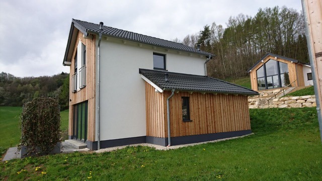 Dwukondygnacyjny dom z białym tynkiem i drewnianą elewacją, z widocznym balkonem i drugim, mniejszym budynkiem z dużym oknem na wzgórzu w tle.