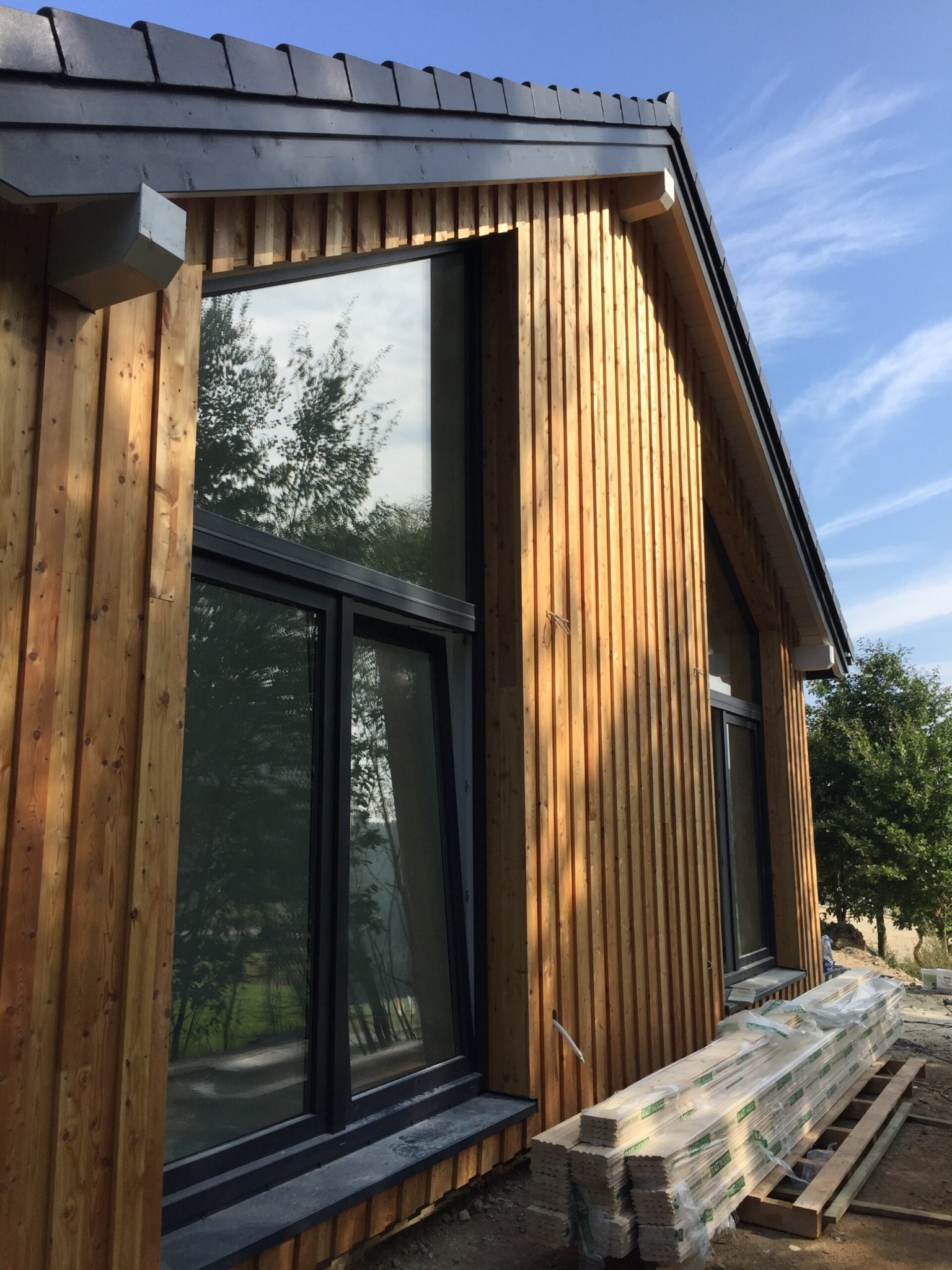 Częściowy widok zewnętrzny domu modułowego z drewnianą elewacją w pionowe deski, czarnymi oknami i stosem drewna budowlanego obok.