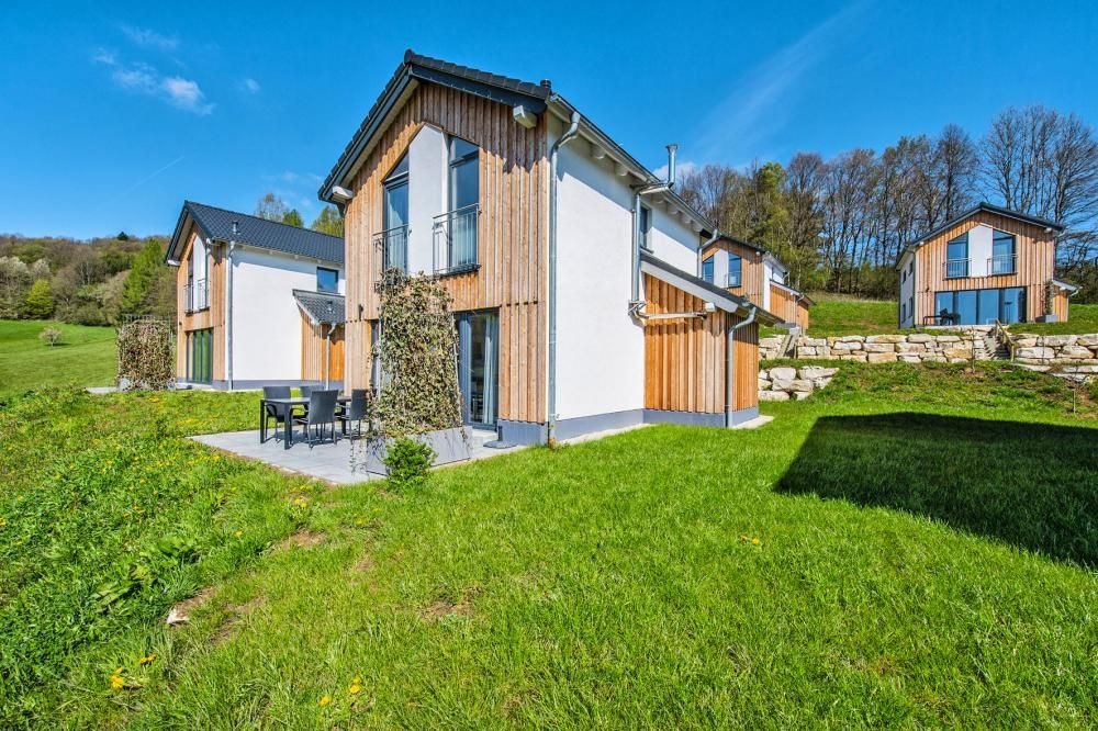 Zestaw kilku nowoczesnych domów z drewnianą elewacją w Opolu, z tarasem i zielonym otoczeniem, w tle wzgórza.