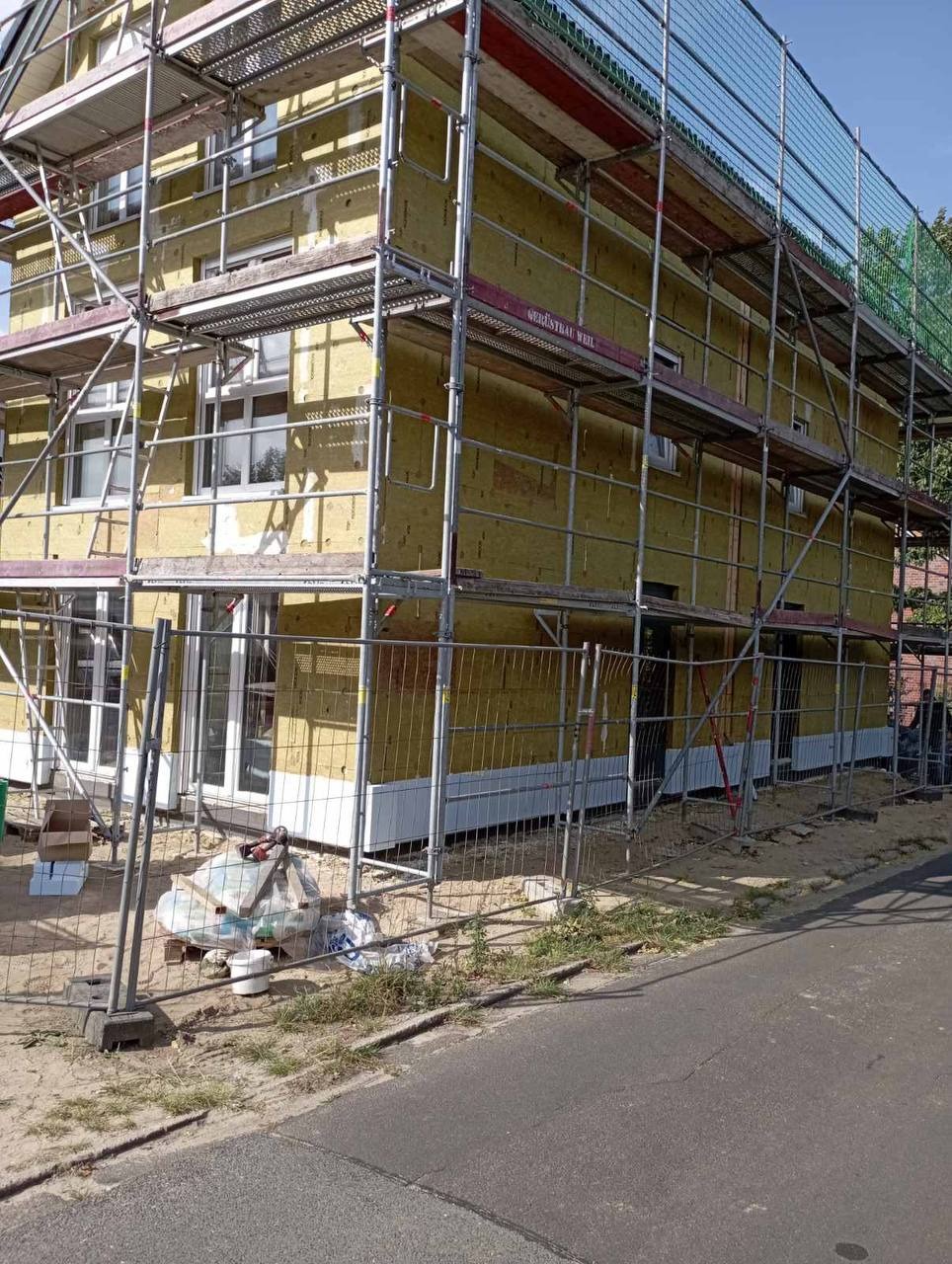 Budynek w trakcie ocieplania, widoczne rusztowania, izolacja z wełny mineralnej na elewacji, częściowo zamontowane białe panele elewacyjne na dole, teren budowy zabezpieczony siatką.