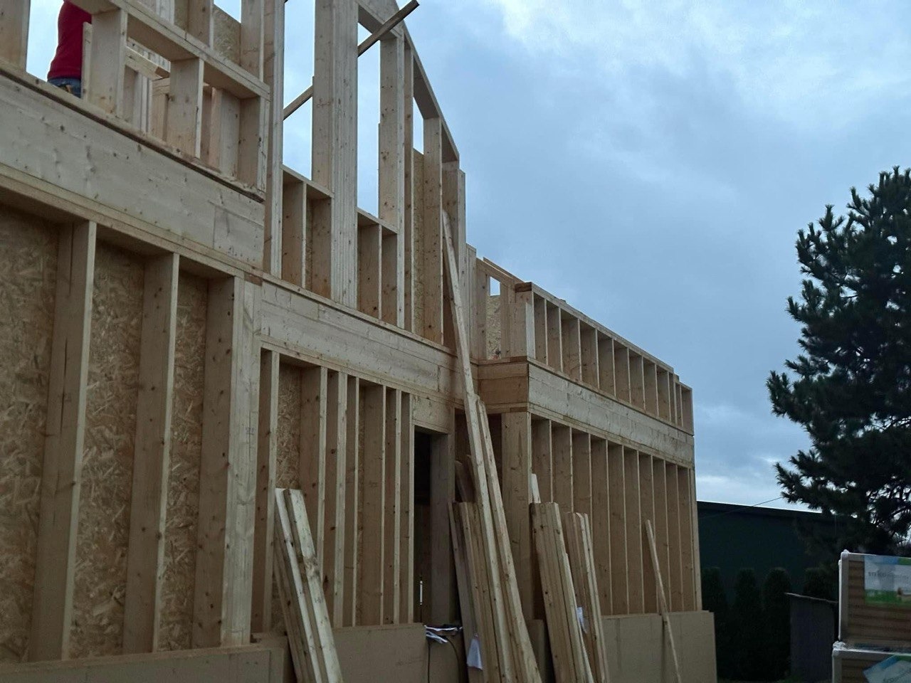 Konstrukcja szkieletowa domu w trakcie budowy, widoczne drewniane elementy ścian i stropów, pochmurne niebo w tle.