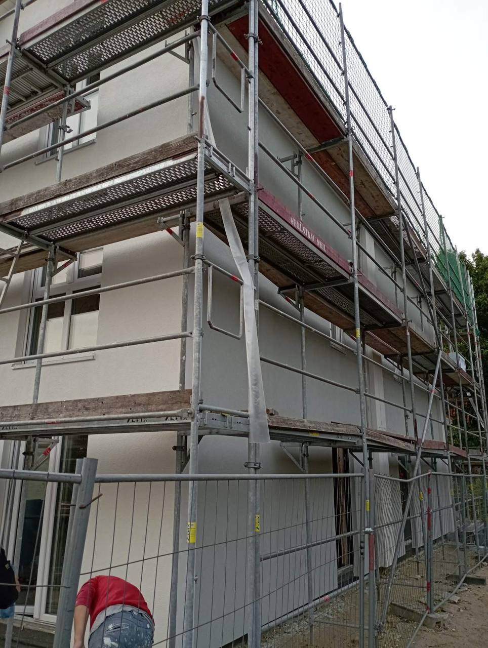 Elewacja budynku w trakcie ocieplania, widoczne rusztowania i siatka zabezpieczająca, pracownik w czerwonej koszulce przy ogrodzeniu.