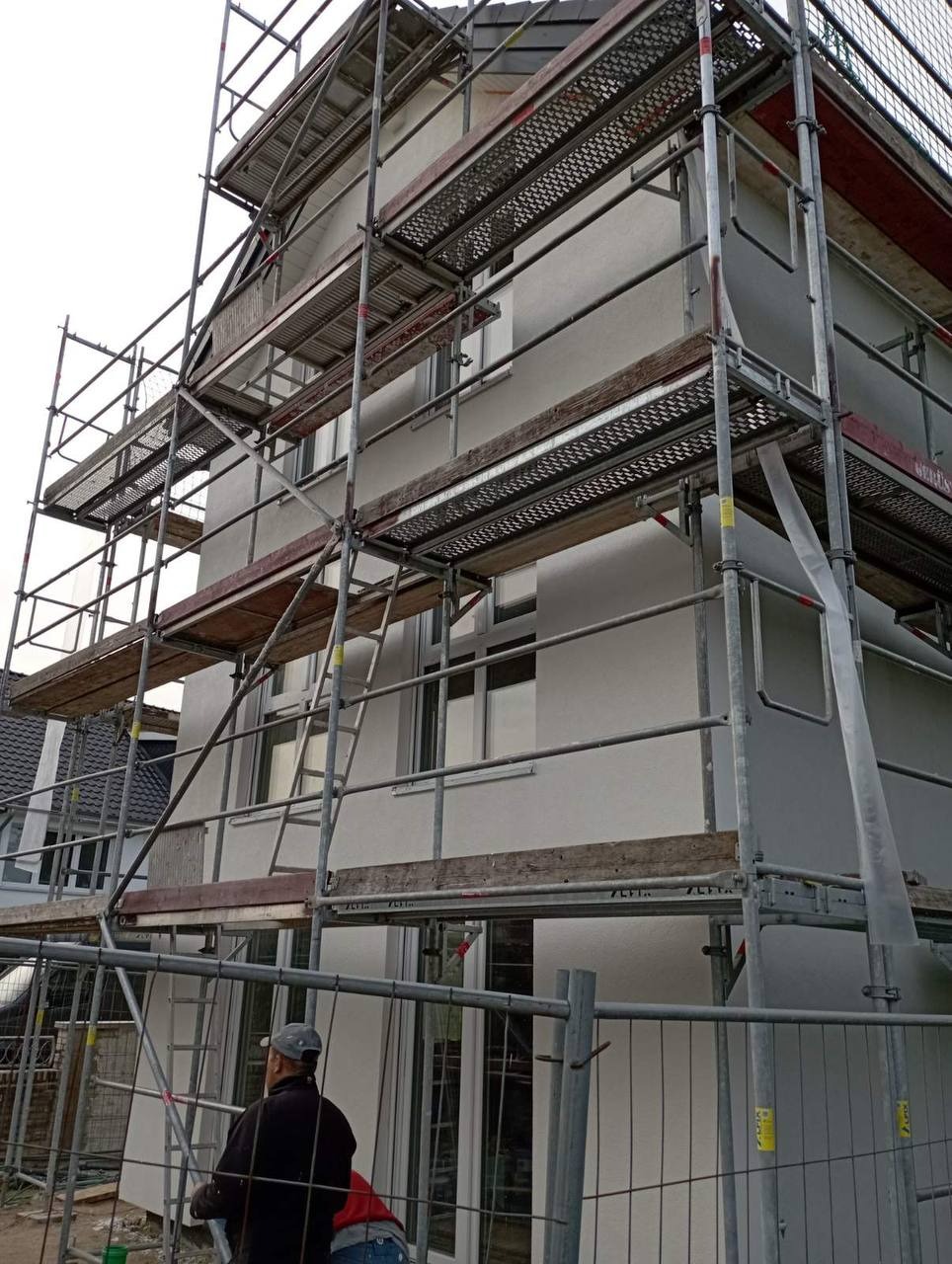 Budowa domu: elewacja budynku z szarym tynkiem w trakcie prac wykończeniowych, widoczne rusztowania i pracownik w czapce w dolnej części kadru.