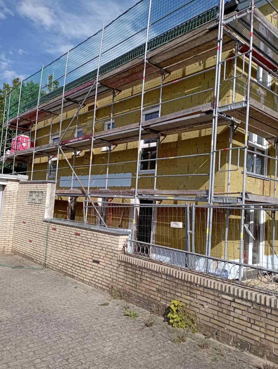 Budynek w trakcie ocieplania styropianem, otoczony rusztowaniem i zabezpieczony siatką ochronną, widoczny ceglany murek i chodnik.