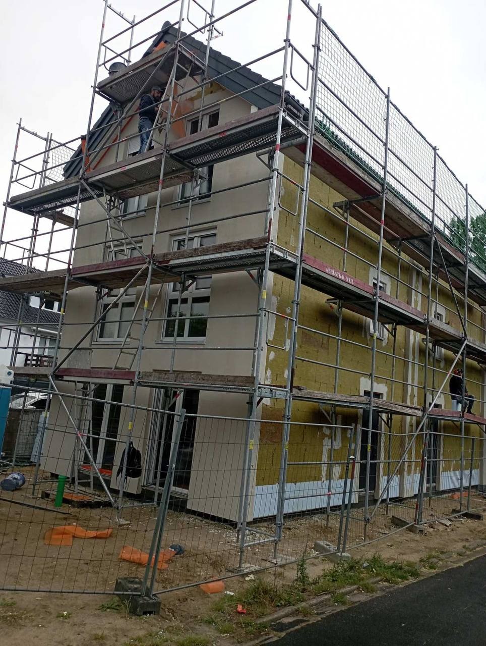 Dwukondygnacyjny dom w trakcie ocieplania elewacji, otoczony rusztowaniem i siatką zabezpieczającą, widoczni robotnicy na rusztowaniu.