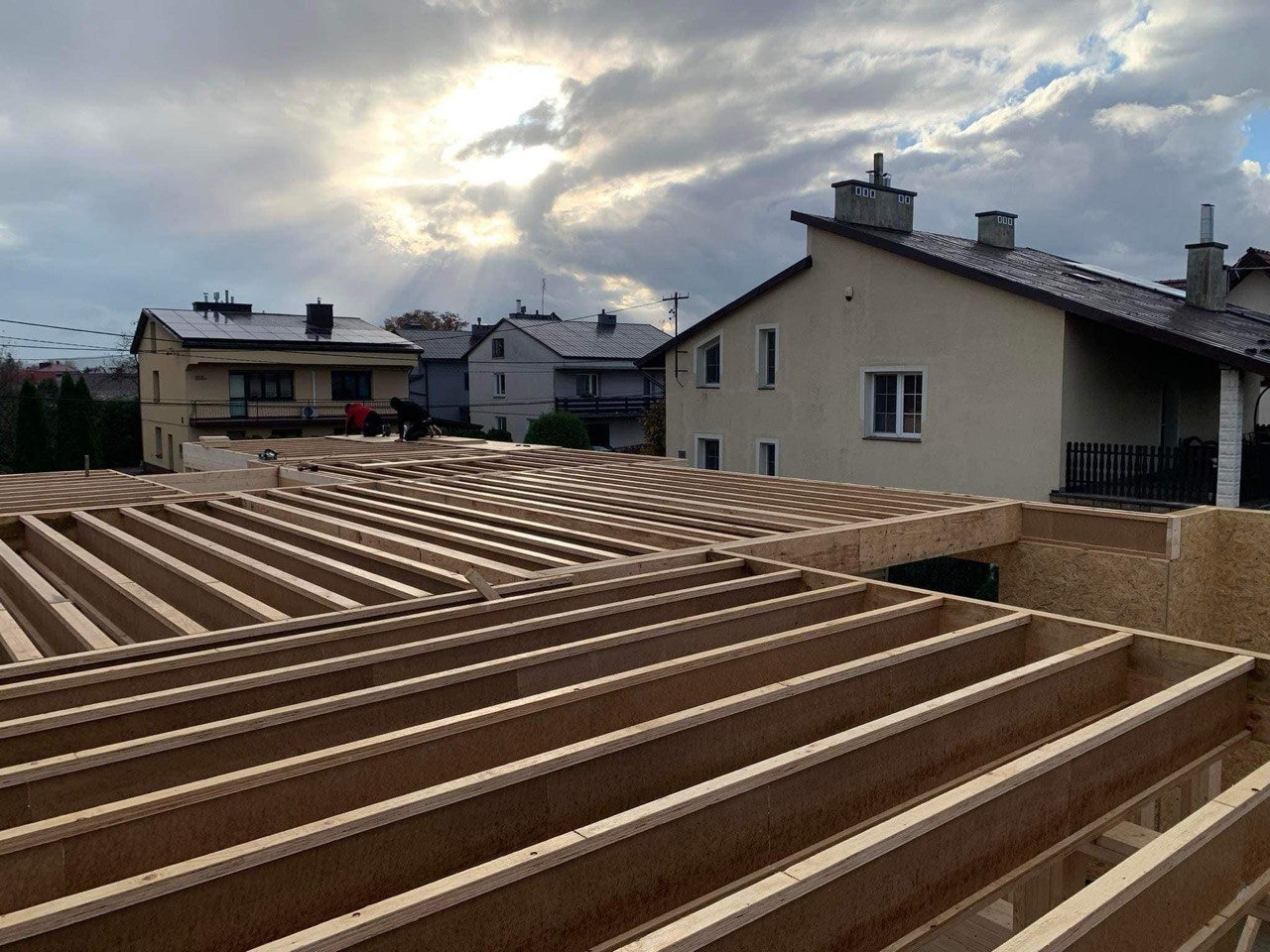 Konstrukcja drewniana stropu w trakcie budowy domu, widoczne belki i płyty OSB, w tle budynki mieszkalne i zachmurzone niebo z promieniami słonecznymi.