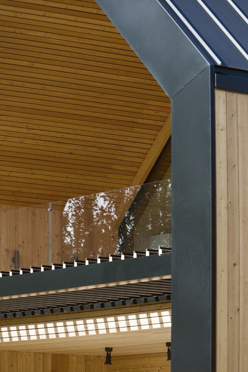 Fragment domu z drewnianą elewacją, widoczny przeszklony balkon z metalowymi wspornikami i drewniany sufit z belkami, oświetlony naturalnym światłem.