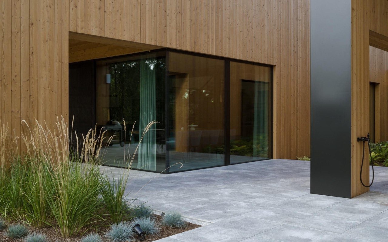 Nowoczesny dom modułowy z drewnianą elewacją, dużymi przeszkleniami i minimalistyczną bryłą, otoczony ozdobnymi trawami i szarą kostką brukową.