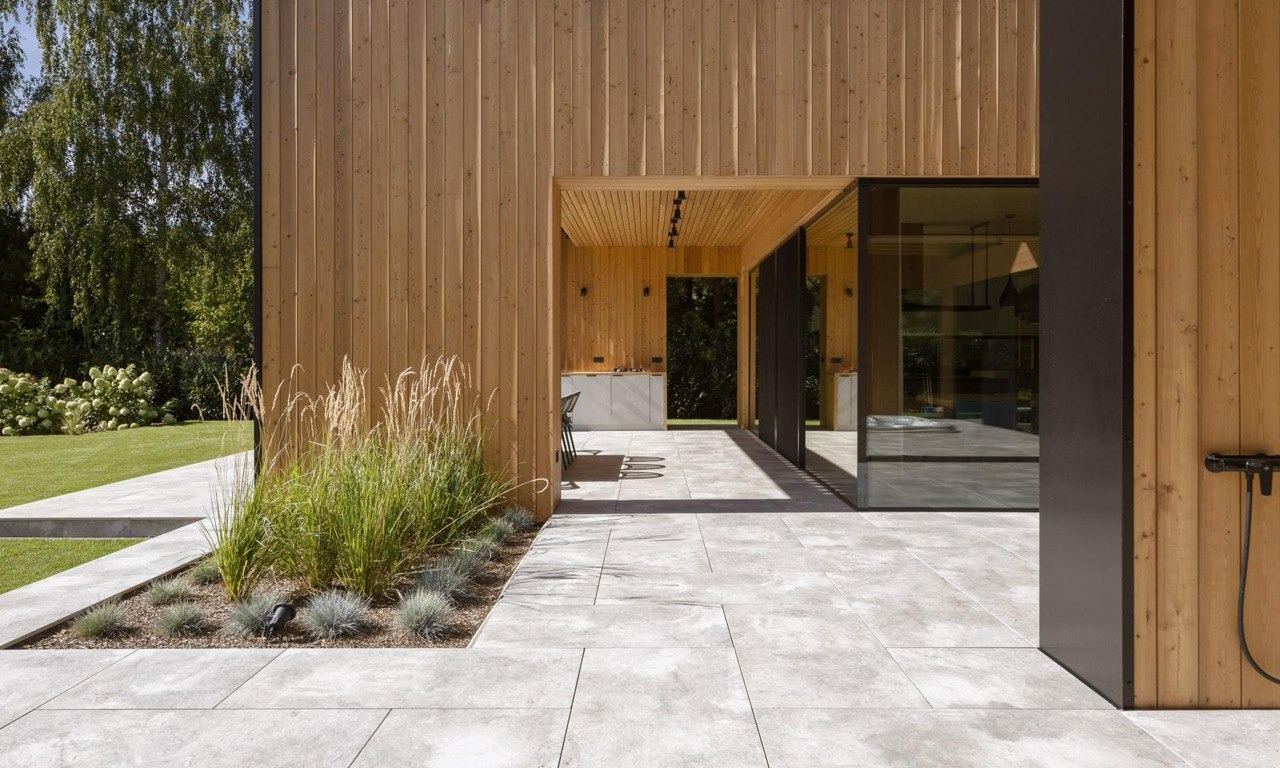 Nowoczesny dom z drewnianą elewacją, dużymi przeszkleniami i minimalistycznym tarasem z szarej kostki brukowej, ozdobiony trawami ozdobnymi.