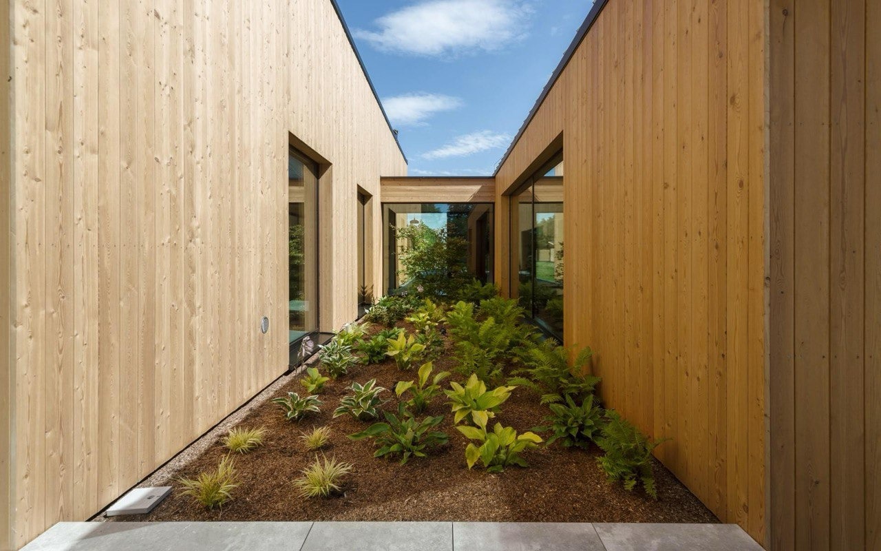 Nowoczesny dom z drewnianą elewacją i wewnętrznym ogrodem, widok na zielone rośliny i niebo przez szklany łącznik.
