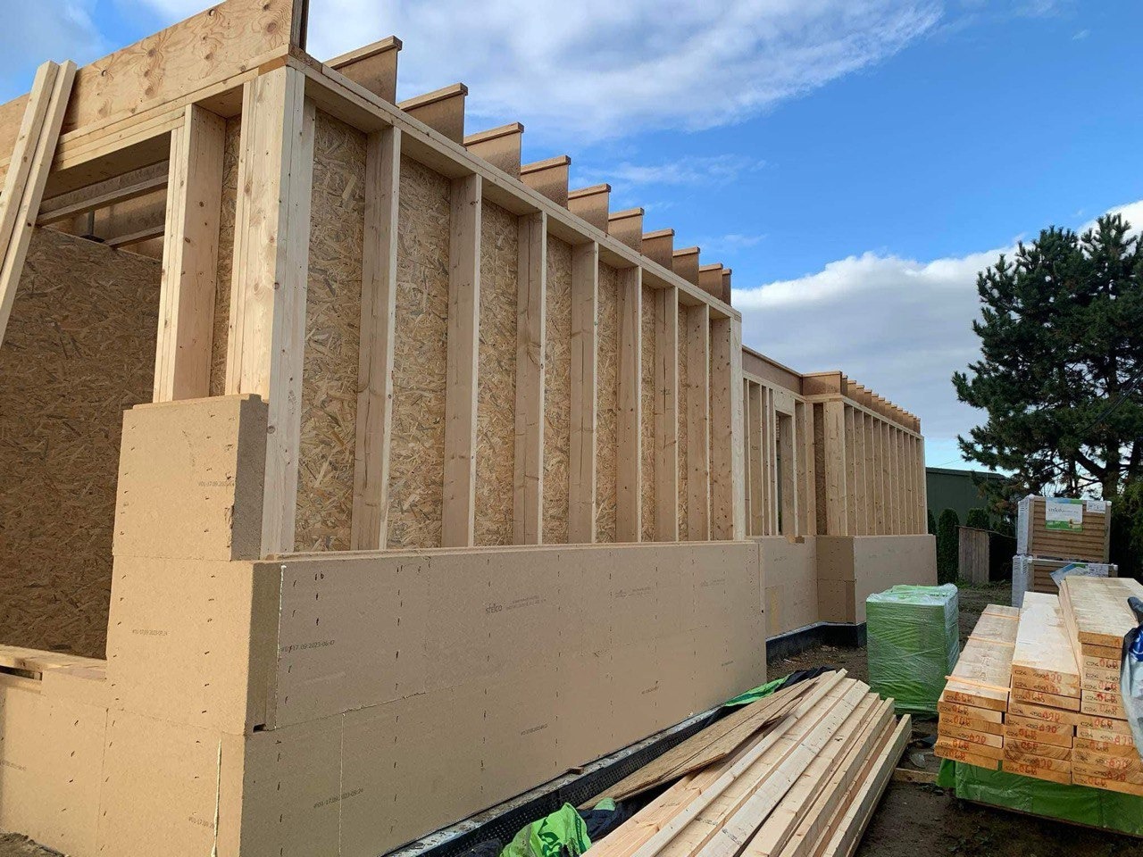 Konstrukcja szkieletowa budynku w trakcie montażu, widoczne elementy drewniane i płyty OSB, materiały budowlane składowane obok, błękitne niebo w tle.