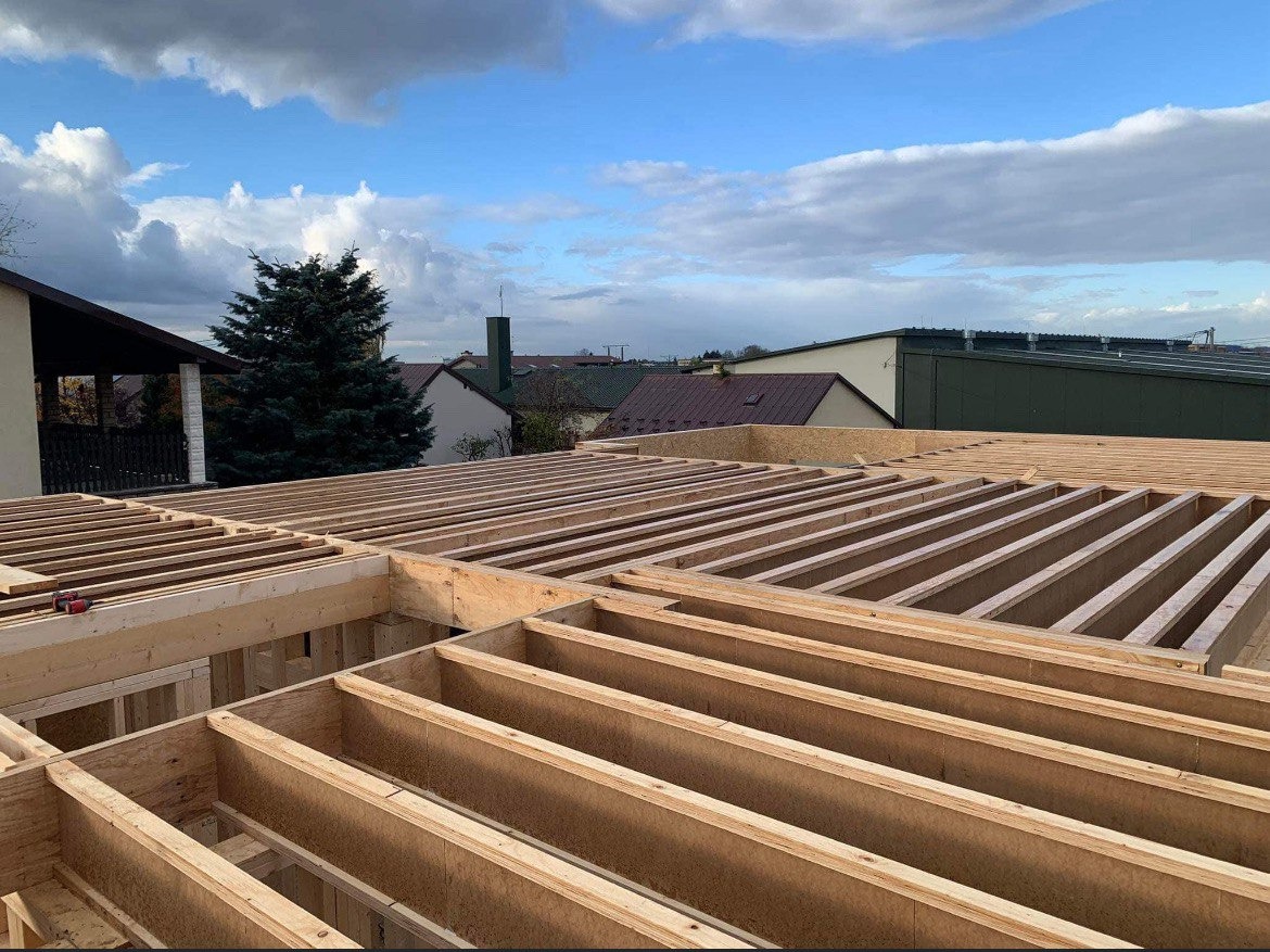 Konstrukcja drewniana stropu budynku w trakcie budowy, widok z góry na belki i płyty, w tle zabudowa miejska i pochmurne niebo.