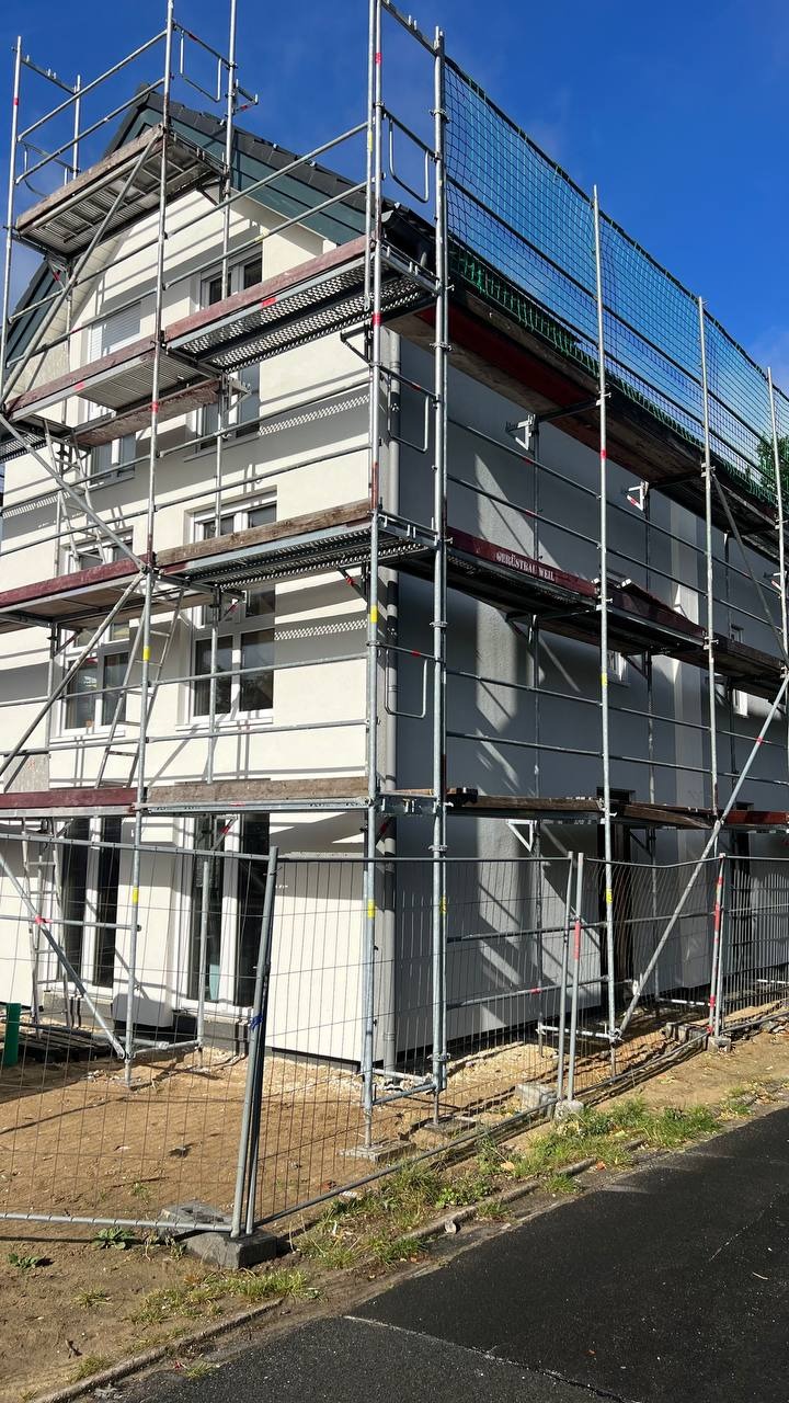 Dwukondygnacyjny dom w trakcie budowy, otoczony rusztowaniami z siatką ochronną, widoczny podział na część w jasnym i ciemnym odcieniu szarości.