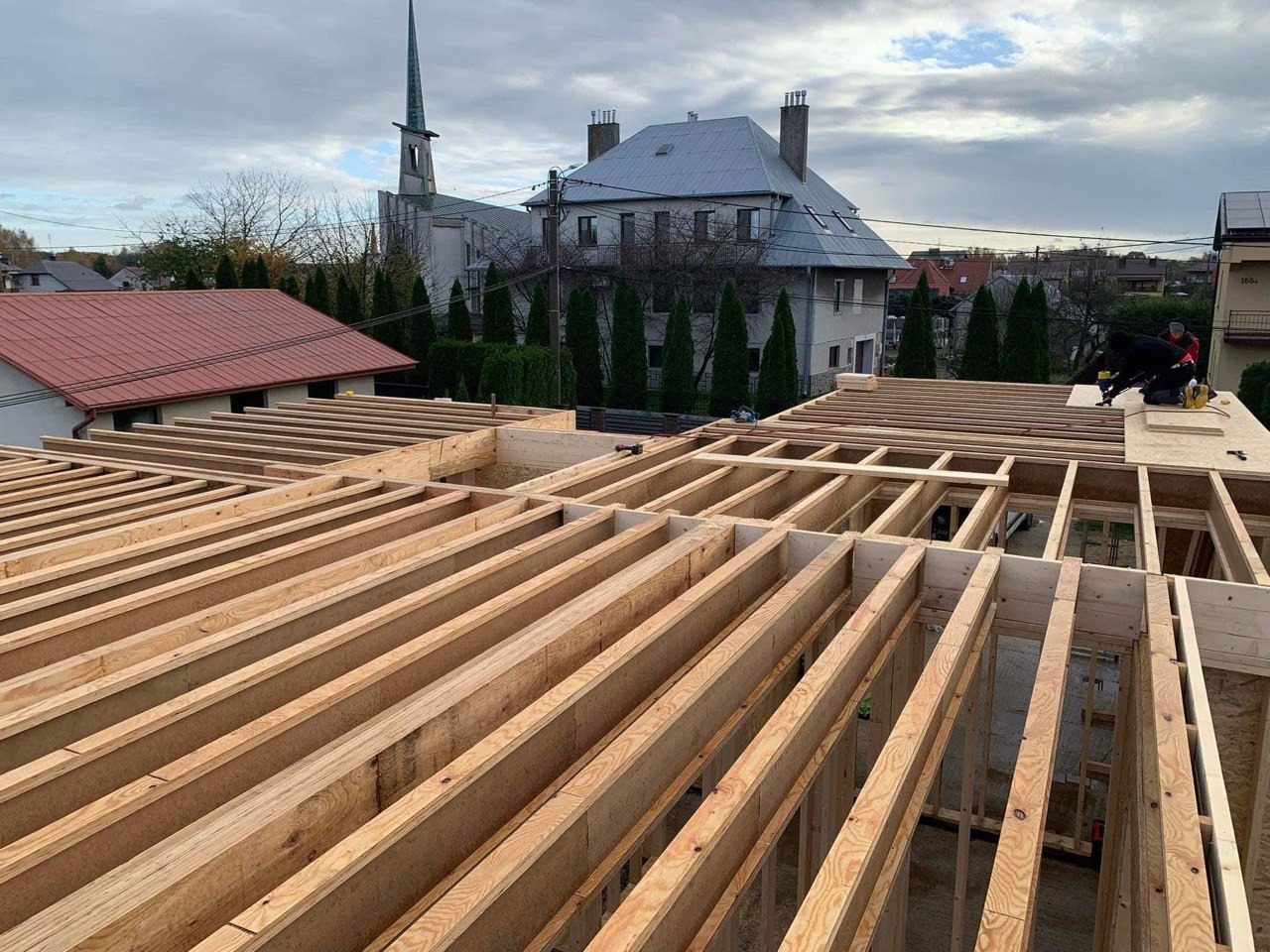 Konstrukcja drewniana szkieletu domu z widocznym kościołem w tle, pracownik na dachu mocuje elementy poszycia.