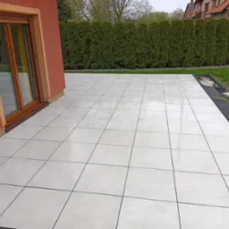 Jasne, kwadratowe płyty tarasowe ułożone przy domu z pomarańczową elewacją i drewnianą stolarką okienną; w tle zielony żywopłot i fragment dachu sąsiedniego budynku.