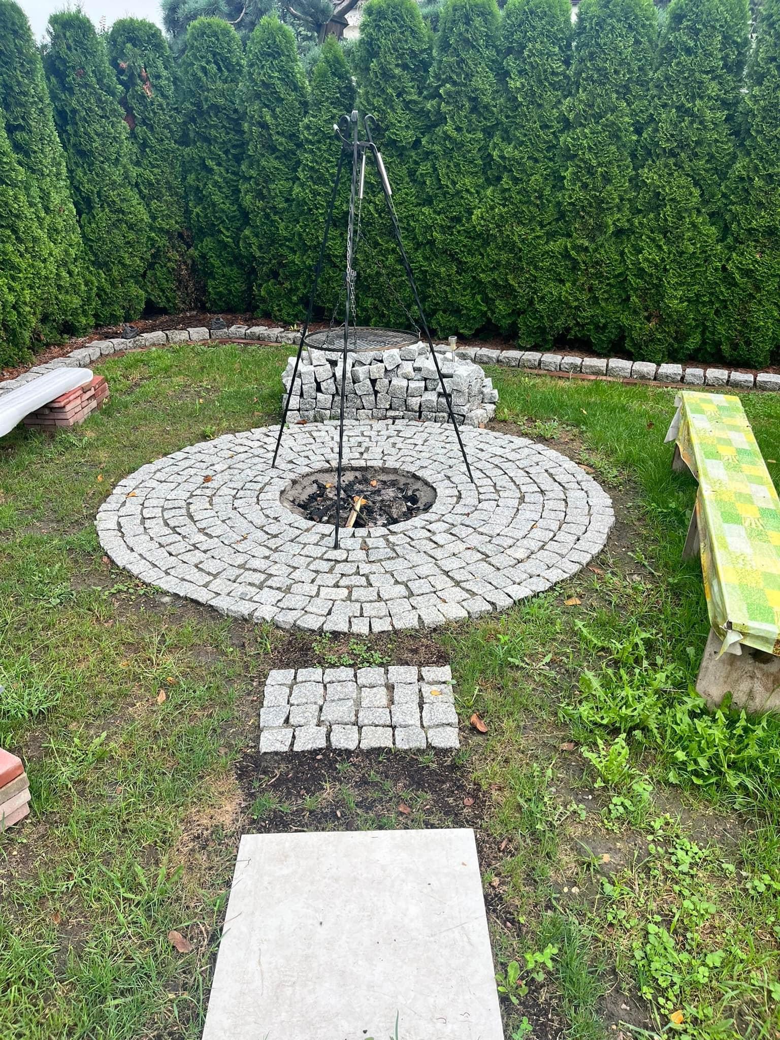 Okrągłe palenisko z jasnoszarej kostki granitowej, otoczone trawnikiem i wysokimi tujami, z trójnogiem nad paleniskiem i ławkami w tle.