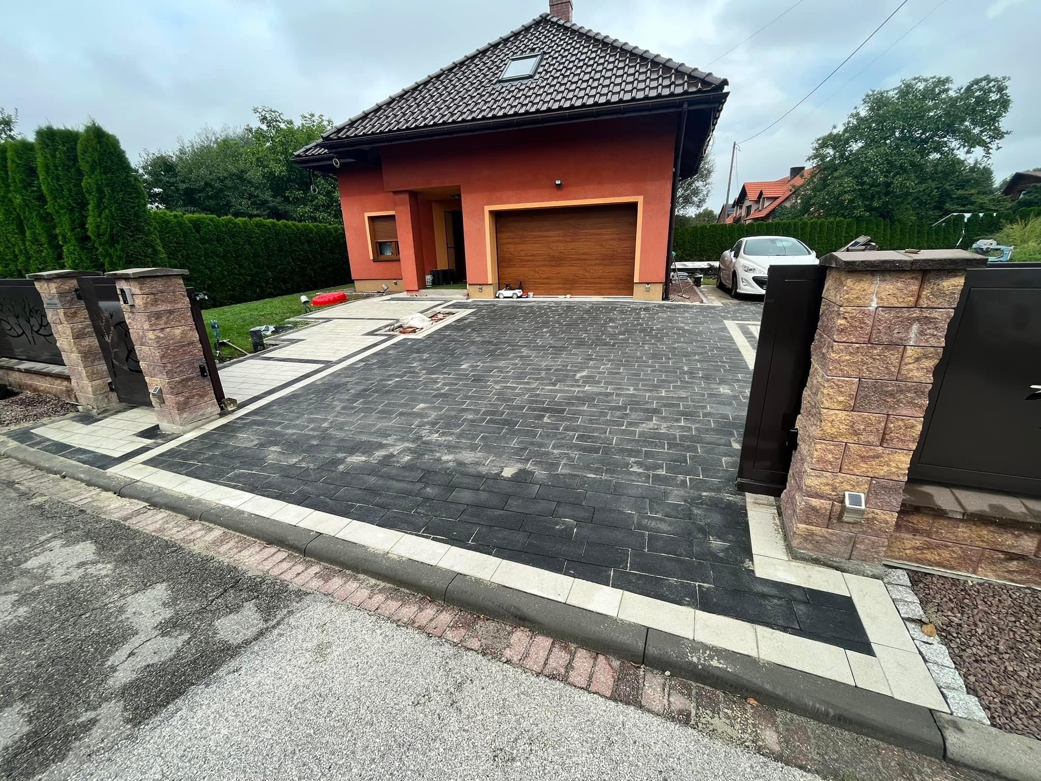 Nowoczesny podjazd z czarnej kostki brukowej obramowany jaśniejszymi elementami, prowadzący do domu z garażem i brązową bramą, całość otoczona zielenią i ogrodzeniem z kamiennymi słupkami.