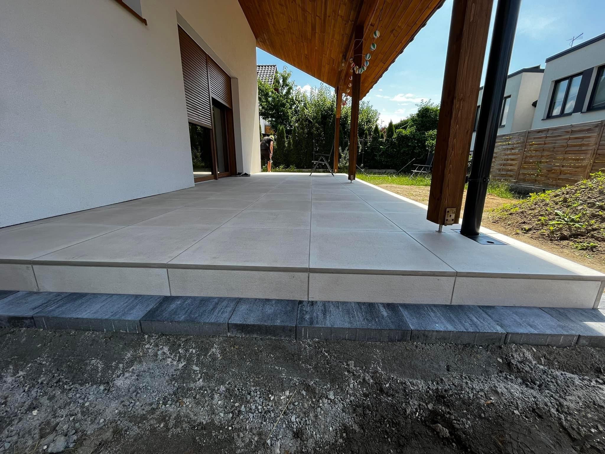 Nowoczesny taras z jasnych, kwadratowych płyt ceramicznych, z ciemnoszarym obrzeżem z kostki brukowej, częściowo wykończony, z widocznym fragmentem ogrodu i elementami konstrukcji z drewna.