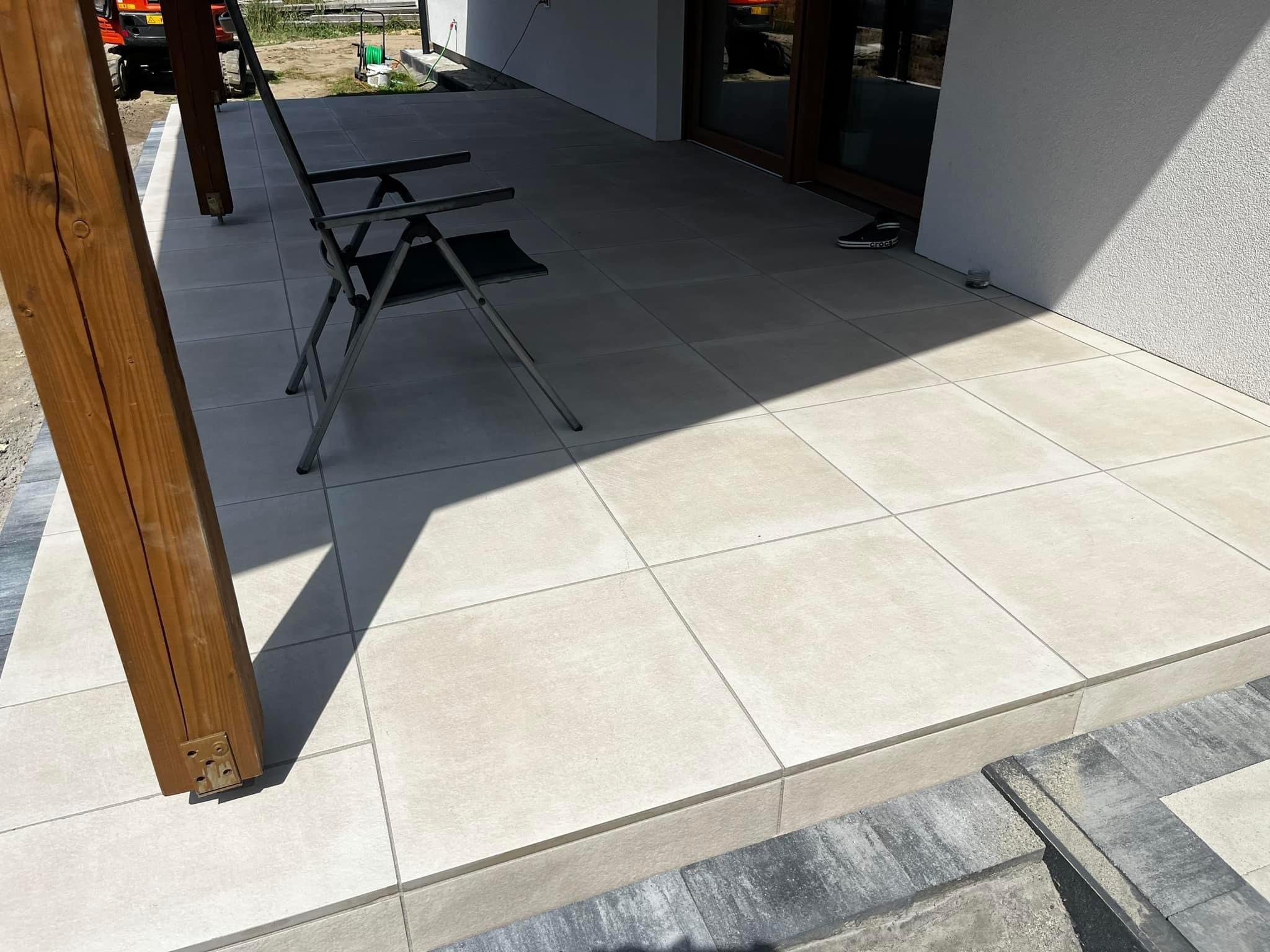 Jasne, kwadratowe płytki tarasowe ułożone na zewnątrz budynku, z widocznym metalowym krzesłem składanym i drewnianym słupem podtrzymującym z metalowymi mocowaniami, z fragmentem kostki brukowej...