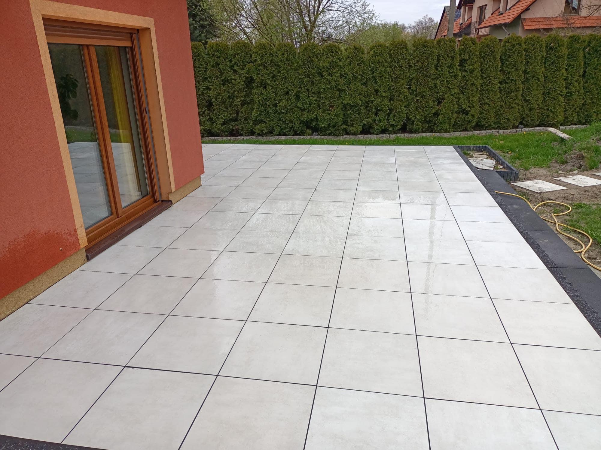 Jasne, kwadratowe płyty tarasowe ułożone przy domu z pomarańczową elewacją i drewnianą stolarką okienną; w tle zielony żywopłot i fragment dachu sąsiedniego budynku.