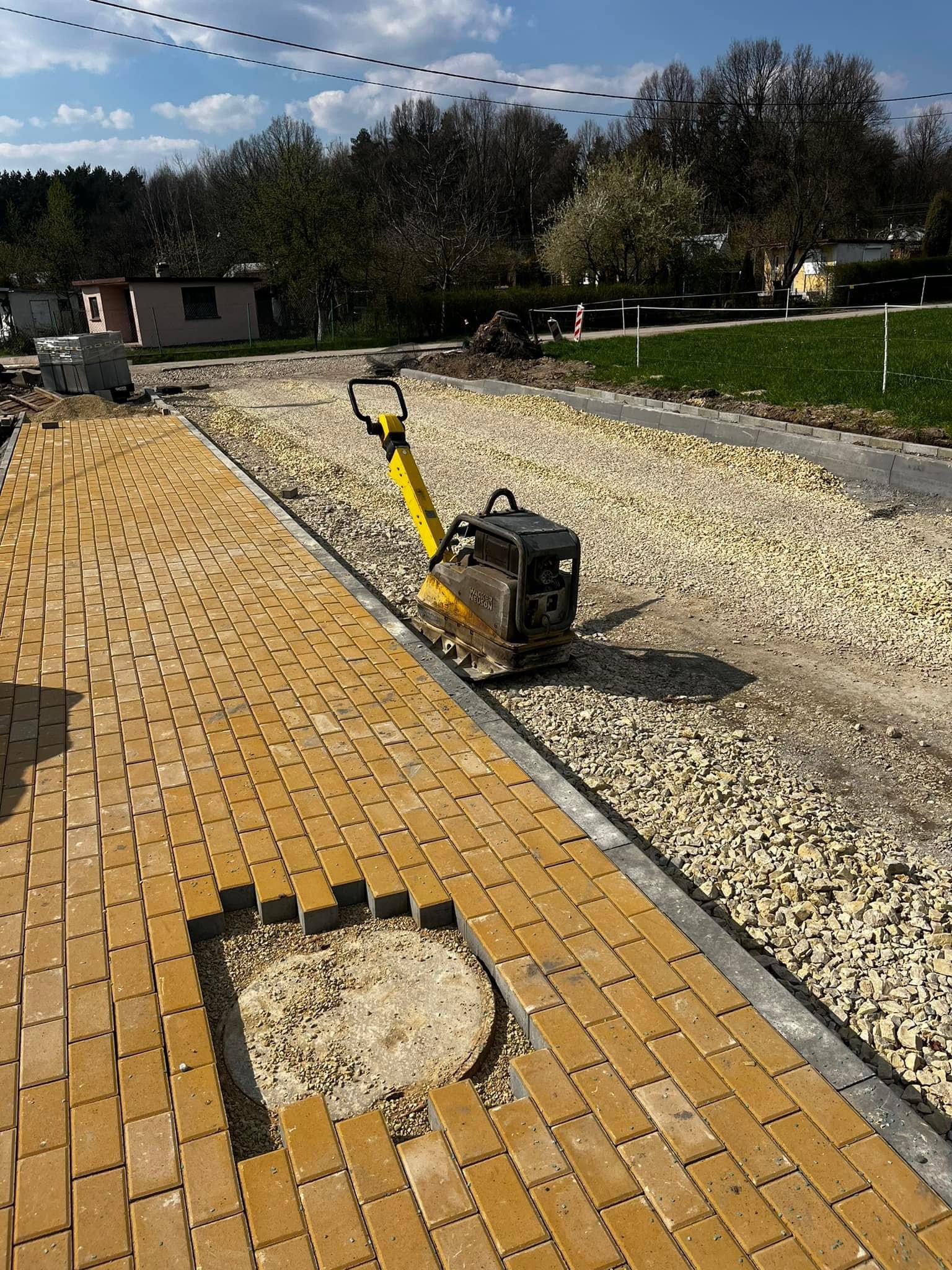 Żółta kostka brukowa w trakcie układania, widoczny otwór rewizyjny oraz zagęszczarka w tle, przygotowywane podłoże z kruszywa.