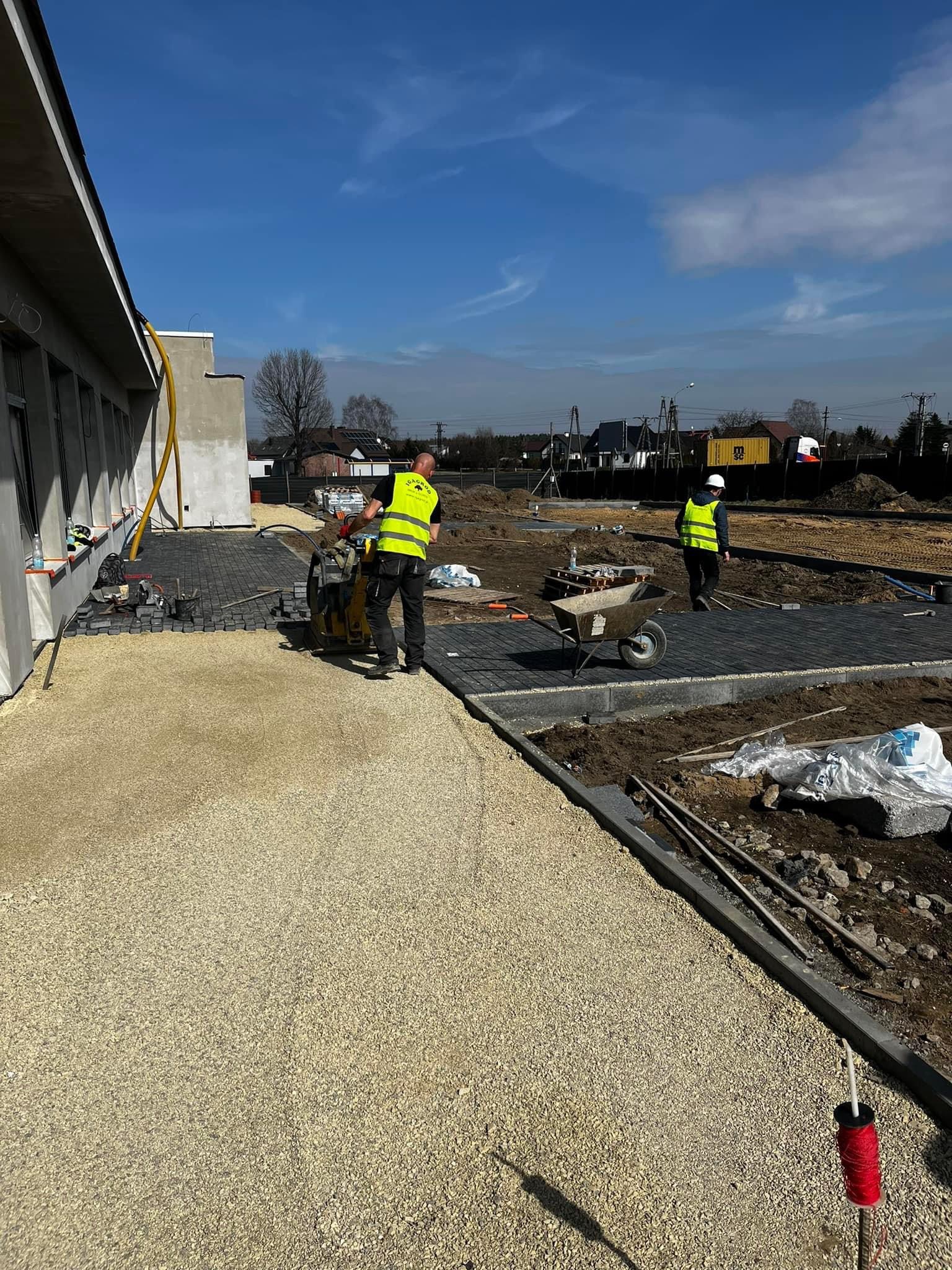 Prace brukarskie w Mikołowie: zagęszczarka wibrująca ubija podłoże pod czarną kostkę brukową, w tle pracownik z taczką i przygotowany teren.